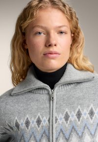 Šedý svetr na zip s vysokým límcem, zdobený diamantovým vzorem v šedé, modré a bílé, vyrobený z měkkého, texturovaného materiálu.
