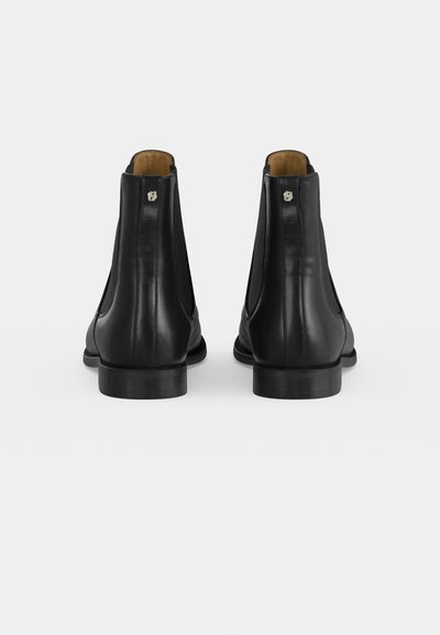 Paire de bottines en cuir noir vues de dos avec des talons bas et un petit détail de logo métallique près du haut.