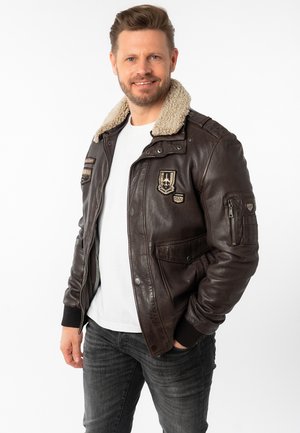Veste en cuir marron avec un col en shearling, plusieurs écussons et une poche zippée. Associée à un t-shirt blanc simple et un jean foncé.