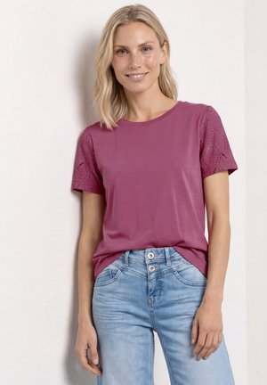 Hymyilevä nainen, jolla on olkapäille ulottuvat vaaleat hiukset, pukeutunut lyhythihaiseen malvanväriseen toppiin, jossa on pitsihihat, ja vaaleansiniset farkut, seisoo valkoista seinää vasten.