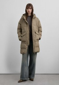 Abrigo acolchado beige con capucha, que cuenta con dos grandes bolsillos delanteros y diseño acolchado. Combinado con vaqueros de pierna ancha desgastados y zapatos de tacón.