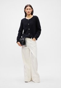 Jūrinės spalvos mygtukinė kardiganas su tekstūriniu paviršiumi, derinama su aukštu liemeniu baltais plačiakojais kelnais. Modelis laiko mažą juodą maišelį su raišteliu.