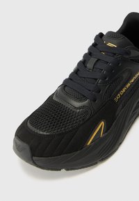 Black sneaker with a mesh and suede upper, featuring gold accents and a textured design. Includes black laces and a padded collar.