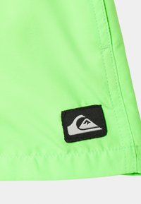 Tela verde neón con textura suave, que presenta un parche negro con un logo blanco en el dobladillo. Se pueden ver los bordes cosidos a lo largo del costado.