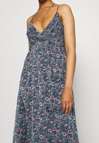 Florale maxi-jurk met blauwe, groene en paarse patronen, verstelbare spaghettibandjes en gerimpelde taille. Lichte stof met een vloeiende uitstraling.