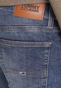 Nahaufnahme von einer blauen Jeans-Rückentasche mit einem Tommy Jeans Lederpatch und einem kleinen Flaggenlogo, kombiniert mit einem beigen Strickpullover.