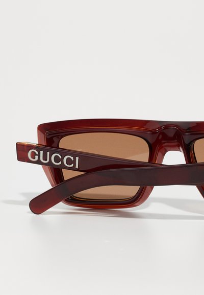 Piros árnyalatú, túlméretes napszemüveg, téglalap alakú lencsékkel, vastag műanyag kerettel és ezüst színű "GUCCI" márkajelzéssel a szárán.