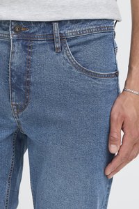 Detailní záběr modrých džín na osobě, která má na ruce stříbrný náramek, světle šedá košile částečně viditelná nad pasem.