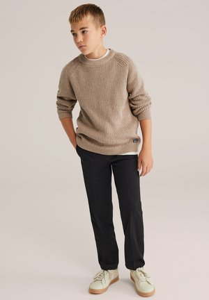 Beige gestrickter Pullover mit geripptem Textur, kombiniert mit schwarzen Hosen und hellen Sneakers, vor einem neutralen Hintergrund stehend.