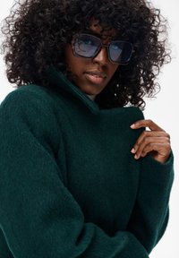 Kobieta z kręconymi włosami, w dużych okularach przeciwsłonecznych i ciemnozielonym, puszystym płaszczu, trzymająca kołnierz płaszcza blisko twarzy.