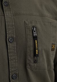 Gros plan de la poche de chemise vert olive avec fermeture éclair noire et logo PME Legend jaune sur la tirette de la fermeture éclair et petite étiquette en tissu.