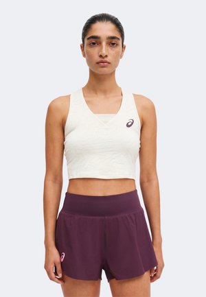 Femme aux cheveux attachés portant un crop top blanc sans manches et un short de sport violet taille haute sur un fond uni.
