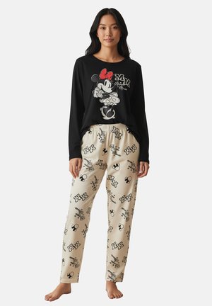Woman wearing black long-sleeve top with Minnie Mouse graphic and beige pants with Minnie-themed logos and text, standing barefoot.