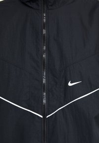 Svart Nike-jakke med glidelås, med hvite detaljer og strukturert stoff. Inkluderer hette og trekantede snittpaneler foran.
