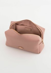 Pink tekstureret kosmetikpose med en buet top, guld lynlås og en cirkulær logo detalje på fronten; indersiden viser et rum.