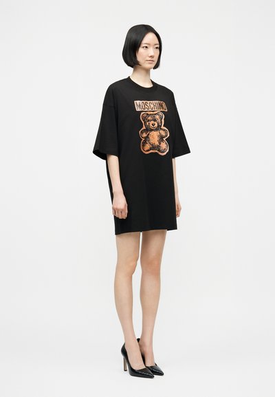 Γυναίκα στέκεται φορώντας μαύρο υπερμεγέθες φόρεμα T-shirt Moschino με πορτοκαλί γραφικό αρκουδάκι και μαύρα ψηλοτάκουνα παπούτσια σε λευκό φόντο.
