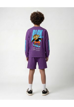 Kind met krullend haar dat een paarse trui en korte broek draagt, met een tekenfilmbeer die skateboardt en de tekst "Keep trying, keep flying."