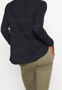 Pull noir en maille avec rayures horizontales texturées, col rond et manches longues, associé à un pantalon ajusté vert olive.