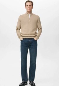 Beżowy dzianinowy sweter z suwakiem przy kołnierzu, w połączeniu z ciemnoniebieskimi prostymi dżinsami. Model ma na sobie czarne buty, ręce w kieszeniach.