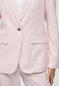 Blazer rose clair ajusté en tissu lisse, doté d'un bouton marron unique, de deux poches avant et d'un col structuré.