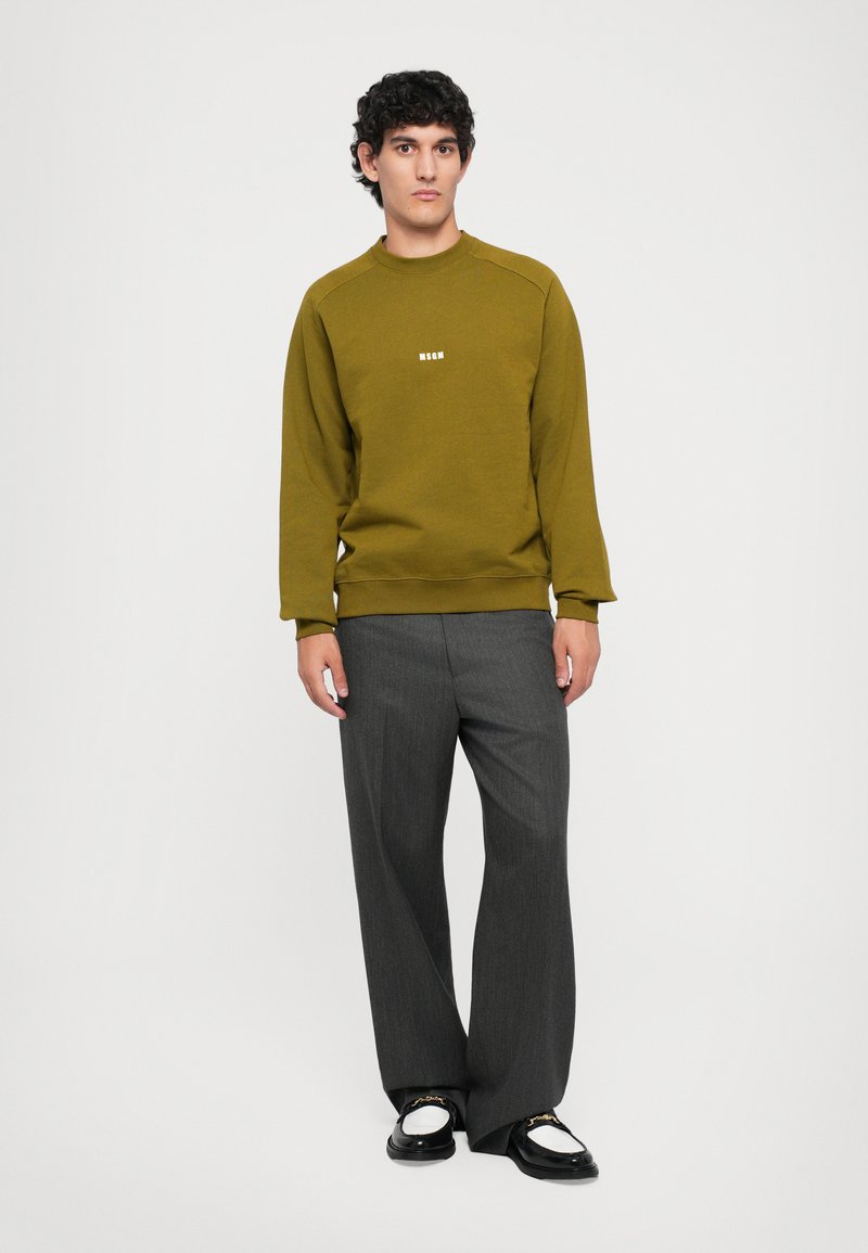 Sweatshirt vert olive avec encolure ras du cou et logo blanc, associé à un pantalon large gris et des chaussures noires et blanches.