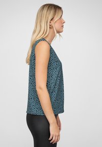 Mouwloze teal top met zwart stippenpatroon, losse pasvorm, lichtgewicht stof en afgeronde zoom, gecombineerd met zwarte leggings.