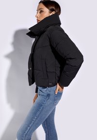Chaqueta acolchada negra corta con cuello alto, textura acolchada y cierre de botones en la parte frontal, que se lleva sobre jeans ajustados azules.