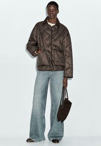 Bruine gewatteerde pufferjas met een kraag, voorzakken en een relaxte pasvorm, gecombineerd met lichtblauwe wijde jeans en een donkerbruine handtas.