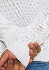 Pull en maille blanche avec un tissage lâche, des poignets en côtes et un motif texturé. Gros plan sur des mains tenant le tissu.