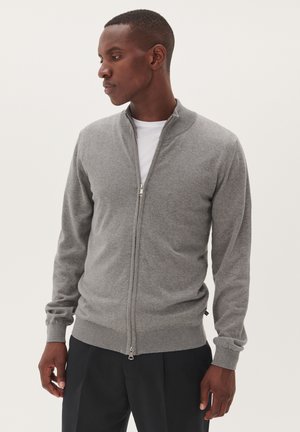 Maglione grigio con zip realizzato in tessuto morbido, con colletto e polsini a coste. Presenta una zip full length e un taglio leggermente aderente, indossato sopra una camicia bianca.