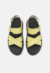 Sandalias amarillas con tiras cruzadas y parte superior texturizada, suelas negras y costuras naranjas. Cuentan con hebillas ajustables para un mejor ajuste y soporte.