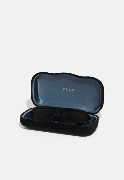 Melni saulesbrilles mīkstā melnā samta maciņā. Iekšpusē ir zila odere ar zelta burtiem "GUCCI". Noapaļoti stūri un gluda tekstūra.