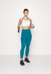 Sport-BH i hvitt, høyt midjekuttet teal treningslegging med sidelomme, og svarte treningssko. Modell som holder motstandsbånd, nøytral bakgrunn.