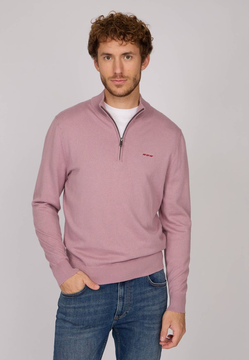 Sweat à zip rose clair avec un col et des poignets côtelés. Présente un petit logo rouge sur la poitrine. Associé avec un jean bleu.