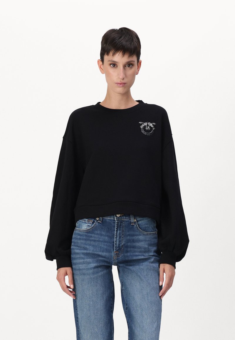 Melna cropped džemperis ar garām bumbieru piedurknēm, ar sudraba logotipa detaļu uz kreisā krūšu. Viegls audums ar gludu tekstūru.