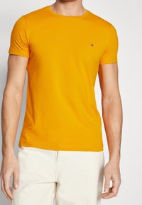 Man som bär en figursydd senapsgul kortärmad t-shirt och off-white byxor, med en liten rektangulär logotyp på vänster bröst.