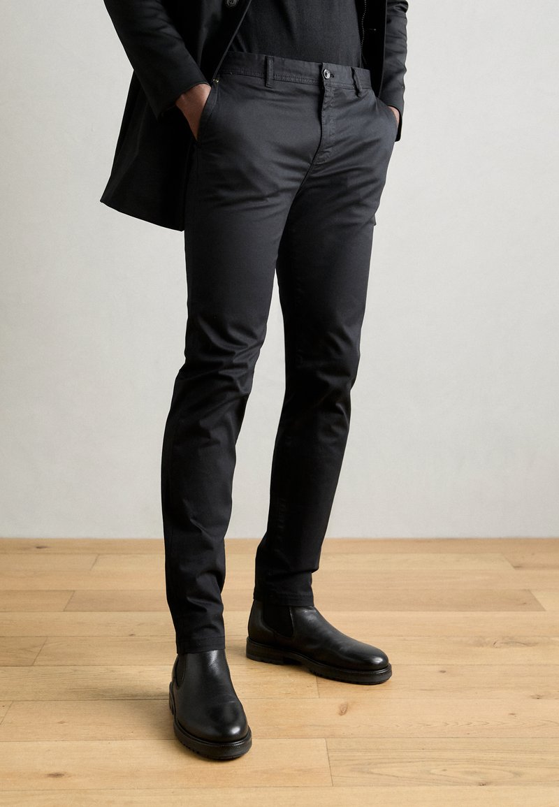 Mustad slim fit chinos, valmistatud siledast kangast. Kombineeritud mustade pahkluu saapastega, eksponeeritud puidust põrandal.