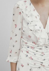 Robe florale blanche avec encolure et manches volantées. Présente de petites fleurs roses et bleues, un tissu froncé pour un bon ajustement et une texture légère.