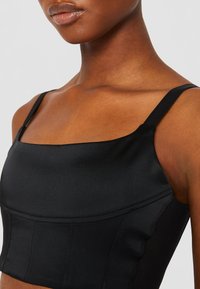Crop top en satin noir avec décolleté carré, fines bretelles et coutures décoratives. Le tissu a une texture lisse avec un léger brillant.