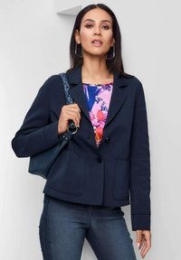 Marineblazer mit zwei Fronttaschen und Knöpfen, über einem blumigen Oberteil in pinken und blauen Farbtönen, kombiniert mit Jeans. Inklusive Handtasche.