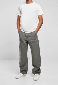 Hombre vestido con una camiseta blanca sencilla, pantalones cargo grises con grandes bolsillos y zapatillas blancas, de pie con las manos en los bolsillos.