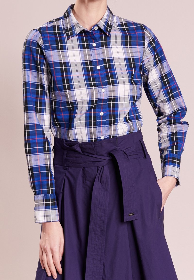 Chemise à carreaux bleue, blanche et noire boutonnée, rentrée dans une jupe taille haute violet foncé avec une ceinture nouée et une main dans la poche.