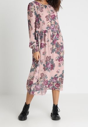 Floral Midikleid in zartem Rosa mit langen Ärmeln, ausgestattet mit einer gerafften Taille und verschiedenen farbigen Blumen. Kombiniert mit schwarzen Stiefeln mit Schnürung.