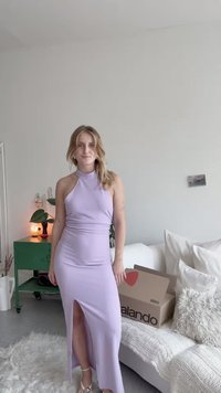 Světle fialové šaty na halter s bočním rozparkem, přiléhavý střih z hladkého materiálu. Na nohách jsou stříbrné sandály na šněrování.