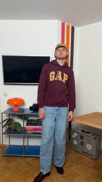 Νέος άνδρας με σκούρο κόκκινο φούτερ GAP και καπέλο στέκεται σε ένα δωμάτιο με τηλεόραση, πορτοκαλί λάμπα, κάμερα, βιβλία και ασημένια βαλίτσα με αυτοκόλλητα.