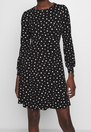 Robe longue noire à manches longues avec des pois blancs, décolleté rond et manches froncées, présentant un corsage ajusté et une jupe évasée.