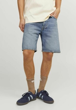 Persoon draagt lichtblauwe spijkershorts, beige sokken met witte patronen en marineblauwe sneakers, staand met één hand in de zak.