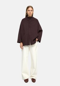 Brązowy wełniany płaszcz z wysokim kołnierzem i zapięciem na trzy guziki, zestawiony z szerokimi białymi spodniami i brązowym obuwiem.