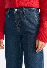 Jeans de mezclilla oscuros de talle alto con bolsillos frontales y costuras en naranja en contraste, combinados con un suéter de punto rojo.