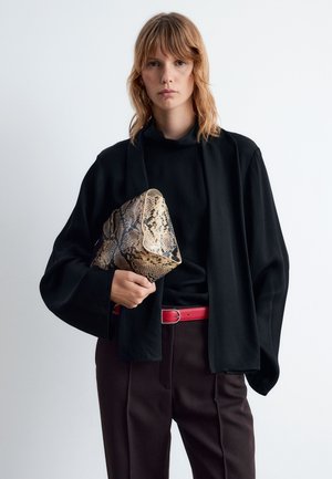 Black loose-fit blouse with a high collar, paired with brown trousers. Holding a textured snakeskin clutch and wearing a red belt.